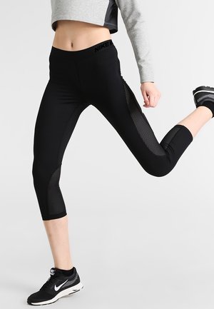 Femme portant des leggings courts noirs Nike avec des panneaux en mesh, des baskets noires Nike, et un haut gris court à manches longues, en posture de course.