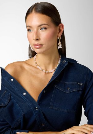 Frau mit blauen Augen und glatten braunen Haaren trägt eine rosa Perlenkette und passende hängende Ohrringe, gekleidet in einem dunkelblauen Denimhemd.