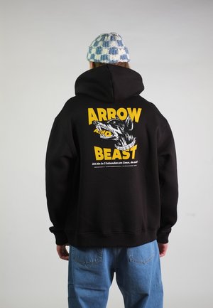 Person trägt schwarzen Hoodie mit gelbem Aufdruck „ARROW AND BEAST“ und aggressivem Hundemotiv, kombiniert mit blauen Jeans und einer karierten Strickmütze.