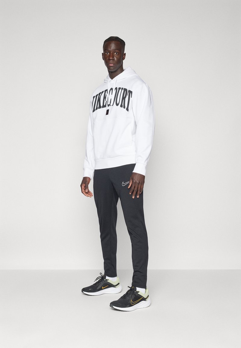 Sudadera blanca con "NIKECOURT" en impresión negra en negrita, combinada con pantalones negros que presentan un pequeño logo de Nike y zapatillas negras con acentos amarillos.