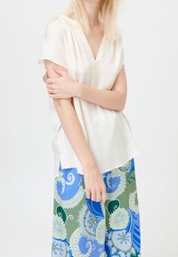 Kvinna i kortärmad vit blus och långkjol med intrikata blommönster i blått, grönt och vitt, poserar mot en enfärgad bakgrund.