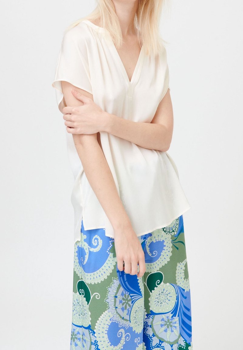 Kvinna i kortärmad vit blus och långkjol med intrikata blommönster i blått, grönt och vitt, poserar mot en enfärgad bakgrund.