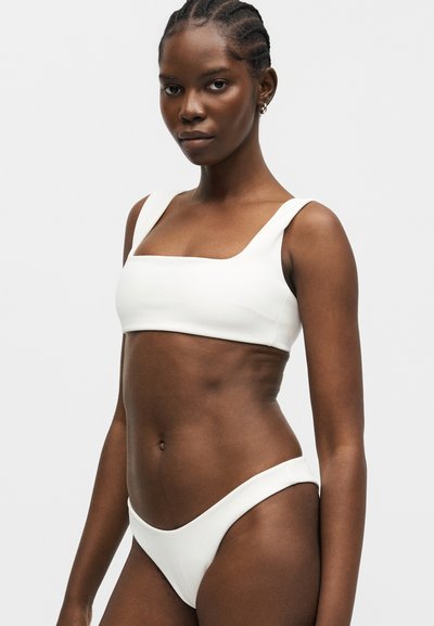 Γυναίκα με πλεξούδες φοράει λευκό μπικίνι τοπ με τετράγωνο ντεκολτέ και ταιριαστό μπικίνι σλιπ με ψηλή μέση, στέκεται μπροστά σε απλό φωτεινό φόντο.