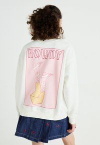Crème sweatshirt met 'HOWDY' op de achterkant, voorzien van een cartoon van laarzen en een gele platformzool. Combineer met een denim rok.