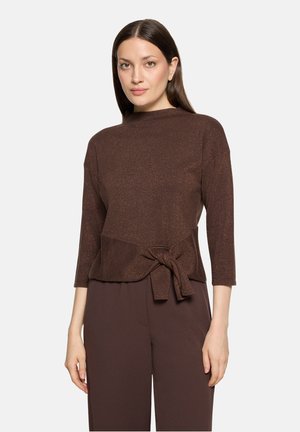 Femme aux cheveux bruns raides portant un haut marron à manches longues avec un nœud à l'avant et un pantalon marron assorti.