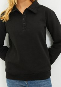 Sudadera negra con cuello camisero, cuatro botones y puños y dobladillo acanalados. Tejido suave sin patrones ni acentos visibles.