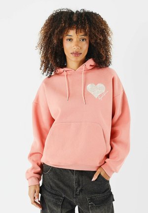 Hoodie rose en tissu doux, doté d'une grande poche avant, d'une capuche avec cordon de serrage et d'un graphique en forme de cœur blanc accompagné d'un détail signature.