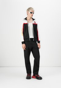Zwart-witte ritsjack met rode accenten, gecombineerd met zwarte cargobroeken en stevige zwart-rode sneakers. Zilveren zonnebril maakt de look compleet.