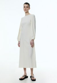 Ecrufarbenes Kleid mit langen, gepufften Ärmeln und gerader Silhouette. Kombiniert mit schwarzen Flip-Flop-Sandalen. Sanfte Textur, minimalistisches Design.
