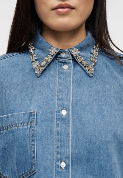 Κοντινό πλάνο μπλε πουκαμίσου από τζιν με γιακά μυτερό διακοσμημένο με κρύσταλλα και λευκά κουμπιά, φορεμένο από άτομο με μακριά σκούρα μαλλιά.