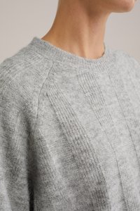 Pelēks adīts džemperis ar ribotu struktūru, ar apaļu izgriezumu un izteiktu paneļu detaļām uz ķermeņa un piedurknēm.