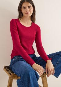 Maglietta rossa a maniche lunghe con scollatura rotonda, abbinata a jeans azzurri a gamba larga. La modella è seduta su uno sgabello di legno con design a listelli.
