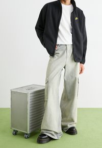 Svart fleece zip-up jacka, vit t-shirt, ljusgrå cargobyxor och svarta platåskor. Metallväska med hjul bredvid. Grön bakgrund.