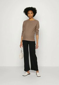 Mulher de pé com cabelo curto e encaracolado, vestindo uma blusa de mangas longas castanha, calças largas pretas, sapatos brancos tipo slip-on e segurando uma pequena bolsa branca.