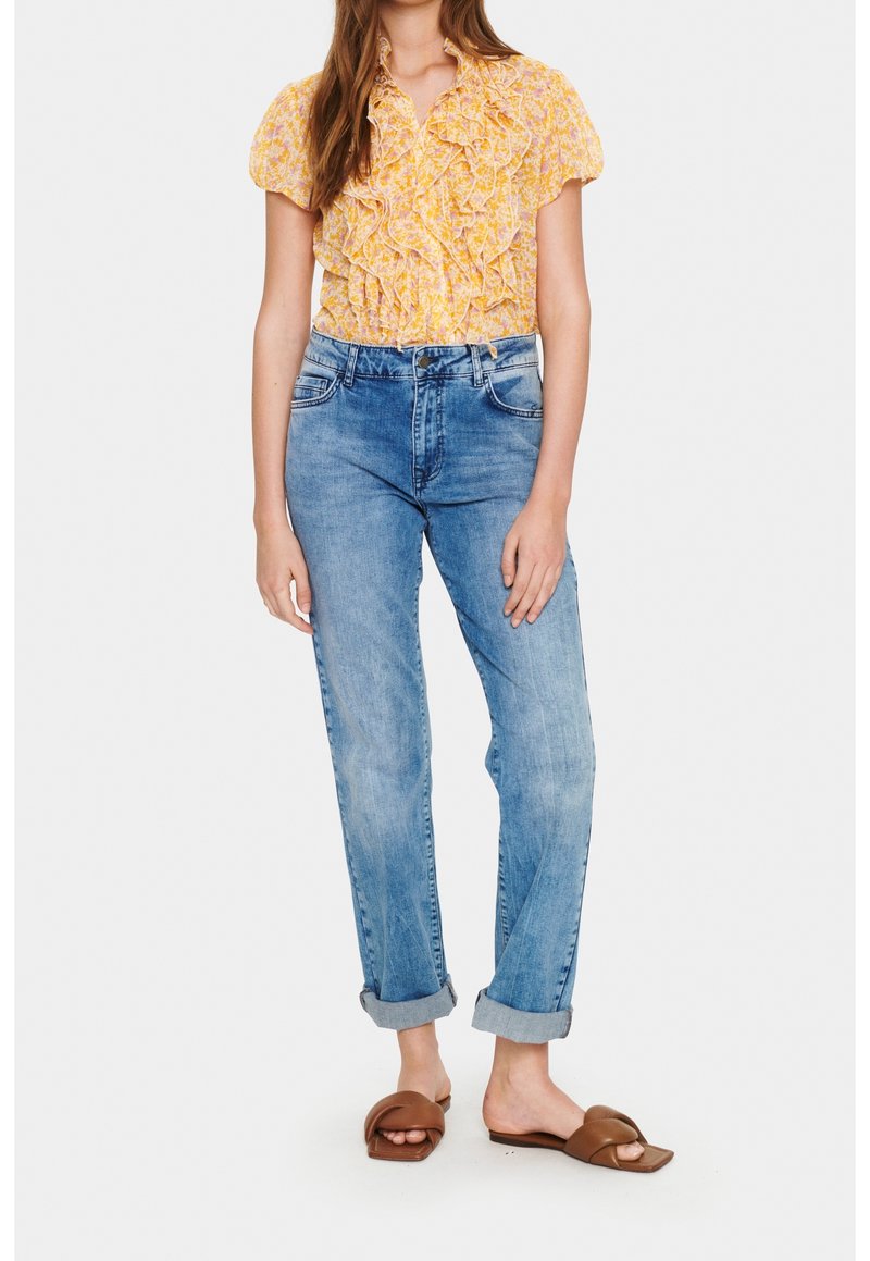 Femme portant un chemisier jaune à fleurs avec des volants, un jean bleu retroussé et des sandales marron à bout ouvert, debout devant un fond uni.