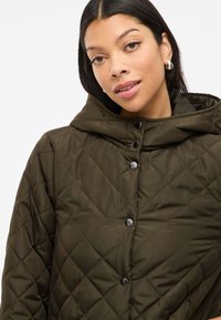 Veste matelassée vert olive avec capuche, dotée de boutons-pression noirs et d'une texture lisse. Présente un design structuré et un matériau durable.