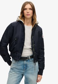 Marine bomberjakke med grå hætte, frontlynlås og sidelommer. Båret over en grå skjorte og lyseblå jeans med et sort bælte.