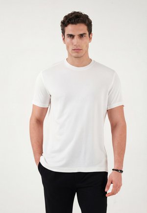 Mężczyzna o ciemnych, kręconych włosach, ubrany w prostą białą koszulkę z krótkim rękawem i czarne spodnie, stojący z jedną ręką w kieszeni na białym tle.