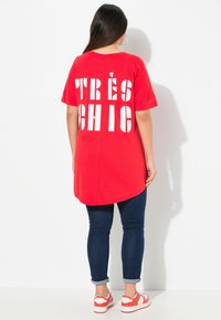 Tunique rouge avec un texte en blanc audacieux "TRÈS CHIC" dans le dos, associée à un jean skinny bleu foncé et des baskets blanches avec des accents rouges.