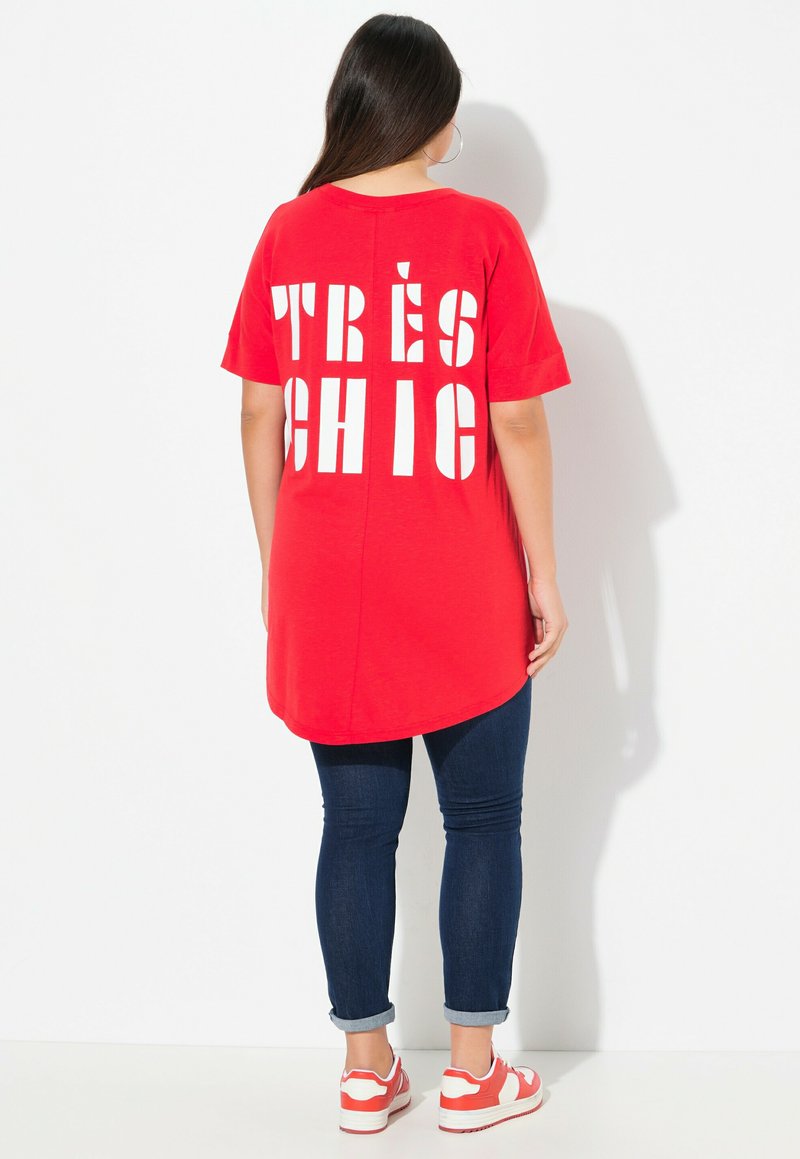 Tunique rouge avec un texte en blanc audacieux "TRÈS CHIC" dans le dos, associée à un jean skinny bleu foncé et des baskets blanches avec des accents rouges.