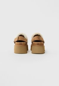 Een paar bruine suède UGG-klompen met een donzige crèmekleurige voering en dikke rubberen zolen, gezien vanaf de achterkant op een witte achtergrond.