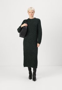 Σκούρο πράσινο, μακρυμάνικο πλεκτό φόρεμα με χαλαρή εφαρμογή, φτιαγμένο από χνουδωτό υλικό. Η μοντέλο κρατά μια μαύρη τσάντα ώμου και φορά γόβες μέχρι το γόνατο.