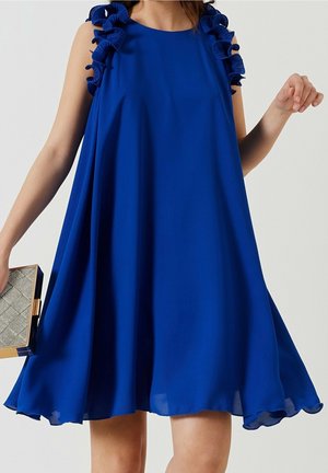 ærmeløs kongeblå kjole med flæser på stropperne og en skørtekant, der flår ud, båret af en person, der holder en lille rektangulær clutch.