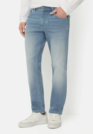 Mand iført lyseblå straight-leg jeans og hvide sneakers, med den ene hånd i lommen, står mod en hvid baggrund.