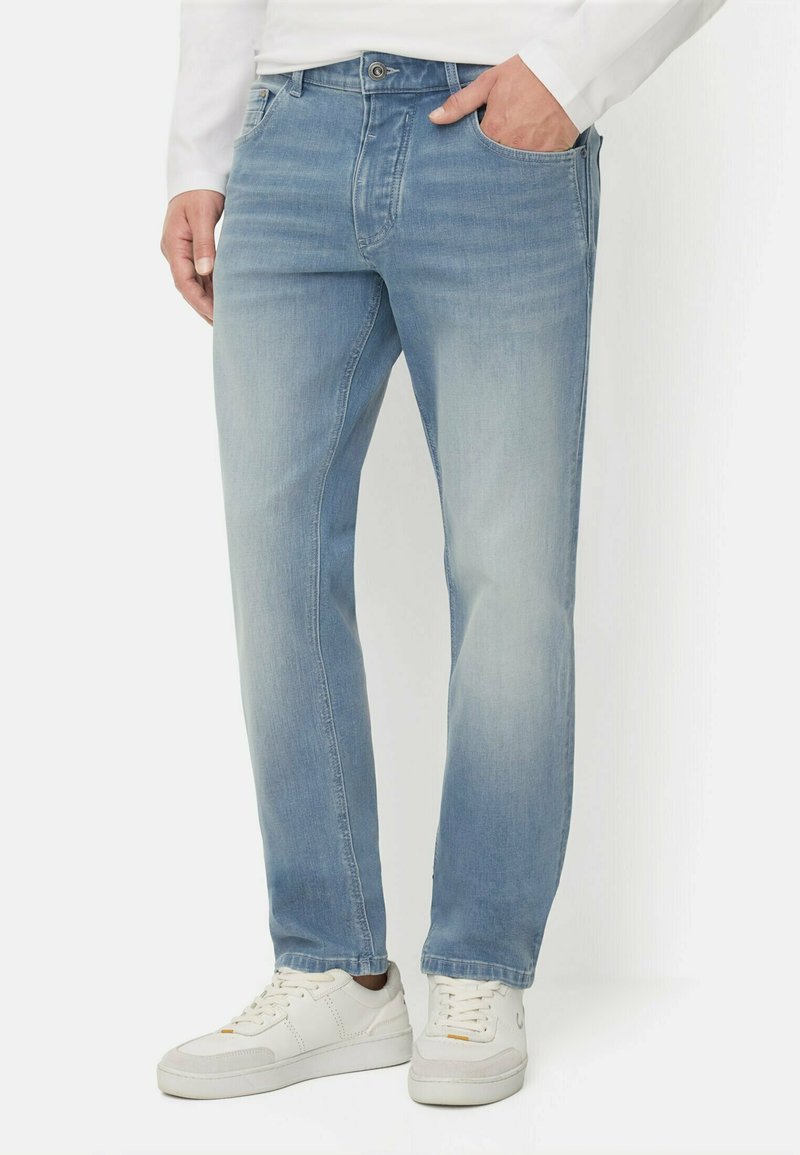 Man wearing light blue straight-leg jeans and white sneakers, with one hand in pocket, standing against a white background.