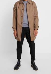 Brązowy, w kratę długi płaszcz z kołnierzem, zapinany na guziki, o teksturze materiału, w połączeniu z szarym swetrem i czarnymi spodniami, dopełniony czarnymi skórzanymi botkami.