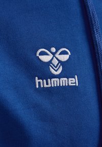 Blått tyg med en slät textur har en vit broderad logotyp som visar en stiliserad biodlare och ordet "hummel."