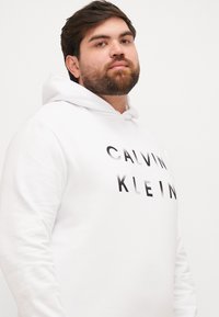 Biała bluza z kapturem z grafiką na froncie, na której widnieją słowa "CALVIN KLEIN" w czarnym, pogrubionym, lekko odbijającym światło stylu. Miękka tekstura, kaptur ze sznurkiem.