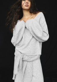 Pulover oversized, de culoare gri deschis, cu mâneci late și decolteu adânc în formă de V, asociat cu o fustă tip wrap, în aceeași nuanță, care are un detaliu cu talie legată.