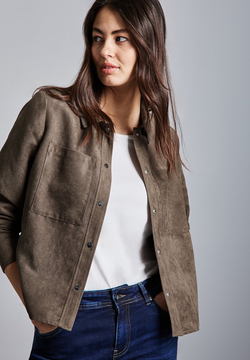 Braune Cordjacke mit strukturiertem Finish, Knopfverschluss, Brusttasche und hochgeschlagenen Ärmeln, getragen über einem weißen Top und blauen Jeans.