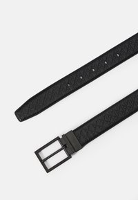 Melnā teksturētā ādas josta ar ģeometrisku rakstu, sudraba taisnstūrveida sprādze un vairāki regulēšanas caurumi gar jostas siksnu.