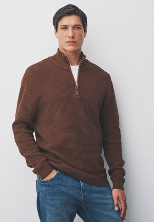 Giovane uomo con capelli scuri che indossa un maglione marrone con mezza cerniera e jeans blu, in piedi con una mano in tasca davanti a un muro bianco.