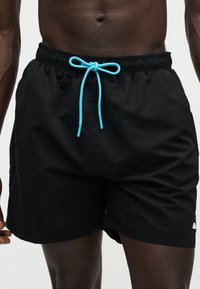 Αρσενικός κορμός με μαύρο μαγιό σορτς και μπλε κορδόνι δεμένο φιόγκο στη μέση, μπροστά σε απλό φόντο.