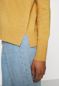 Gros plan d'une personne portant un pull en tricot jaune moutarde avec des poignets côtelés et un jean en denim bleu clair.
