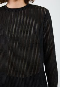 Black long-sleeve top with a textured, grid-like pattern, featuring sheer panels and a round neckline. Soft fabric with a fitted shape.