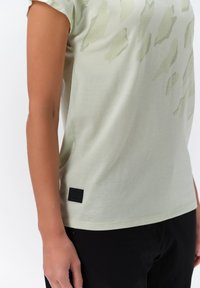 Camiseta de manga corta verde claro, hecha de una tela suave, con un sutil patrón abstracto. Incluye una etiqueta de tela negra en el dobladillo.