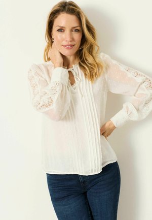 Frau mit welligem blondem Haar, die eine weiße Spitzenbluse und dunkelblaue Jeans trägt, lächelt mit einer Hand am Hals und der anderen an der Hüfte.