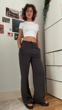 Calças largas cinzentas com pregas frontais e bolsos laterais, combinadas com uma t-shirt curta branca. O ambiente apresenta um piso de madeira clara.