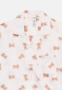 Kortärmad pyjamaströja i vitt tyg prydd med ett mönster av nallar och rosa hjärtan, med en bröstficka och knappar.