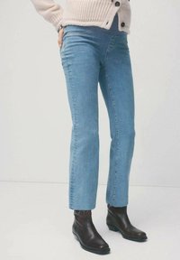 Osoba nosząca jasnoniebieskie jeansy o prostych nogawkach, brązowe botki za kostkę i beżowy sweter na guziki stojąca na tle jednolitego tła.