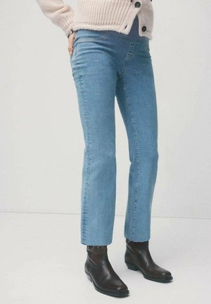 Osoba nosząca jasnoniebieskie jeansy o prostych nogawkach, brązowe botki za kostkę i beżowy sweter na guziki stojąca na tle jednolitego tła.