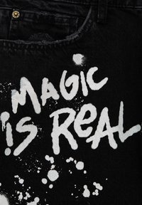 Texte blanc peint à la bombe indiquant "MAGIC IS REAL" avec des éclaboussures de peinture sur du tissu en denim noir près d'une poche et d'un passant de ceinture.