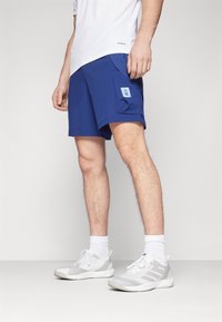 Pantaloni sportivi blu navy con una tasca sulla gamba sinistra, dotati di una toppa logata azzurro chiaro. Abbinati a scarpe da ginnastica bianche e calze.