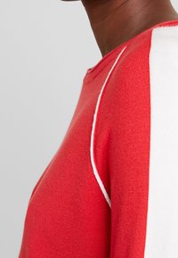 Maglione rosso lavorato a maglia con una striscia bianca lungo la spalla. Caratterizzato da un collo rotondo e una texture morbida, che mette in risalto i colori a contrasto.