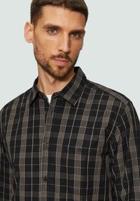 Chemise à carreaux noire et beige, à boutons, avec un col pointu, dotée d'une poche poitrine et d'un tissu texturé. Vue de face de la chemise.