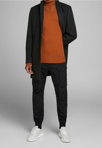 Schwarzer langer Mantel über einem orangefarbenen Rollkragenpullover, kombiniert mit schwarzen Cargo-Hosen und weißen Turnschuhen. Zu den wichtigsten Merkmalen gehören eine lockere Passform und ein geschichtetes Design.